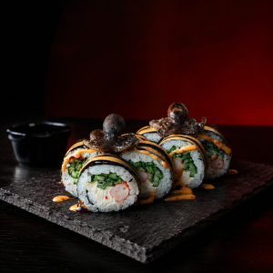 Maki sushi rolls with cucumber and crab, wrapped in nori, topped with octopus and orange spicy mayo on a slate board.